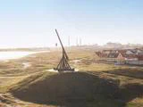 En stor trebuchet af træ står på en græsklædt bakke med udsigt over en kystby med huse og industribygninger i baggrunden under en klar himmel.