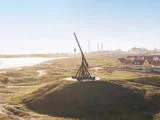 En stor trebuchet af træ står på en græsklædt bakke med udsigt over en kystby med farverige huse og et industriområde i baggrunden.