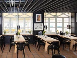 En lys, moderne café med træborde, sorte stole, hængelamper og planter i midten. Store vinduer lukker naturligt lys ind.