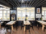 Moderne restaurantinteriør med træborde, sorte stole, hængelamper og store vinduer, der lukker naturligt lys ind; planter og kunstværker pynter i lokalet.