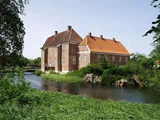 En stor, historisk murstensbygning med røde tegltage ligger ved en rolig flod, omgivet af grønt græs, buske og træer under en klar, blå himmel.