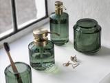 Badeværelsestilbehør i grønt glas med guldfarvede pumper og låg er anbragt i en hvid vindueskarm sammen med en tandbørste, en halskæde og et par øreringe.
