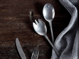 En metalkniv, gaffel, ske og spork ligger på et mørkt træbord ved siden af et foldet grå- og hvidternet klæde.