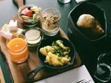 En træplade med røræg, ost i skiver, frugt, granola, juice og brød i en kurv, sat på et bord med redskaber og et glas.