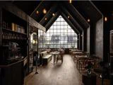 Moderne, svagt oplyst café med træborde og -stole, højt hvælvet loft, store gittervinduer og hængende pendellamper, som skaber en hyggelig og stilfuld atmosfære.