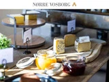 En træplade med forskellige oste og syltetøjsglas står på et bord under Nørre Vosborgs logo med osteetiketter i baggrunden.