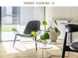 En moderne loungestol og et rundt bord med en potteplante i et solbeskinnet, minimalistisk rum. Teksten "NØRRE VOSBORG" står øverst.