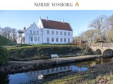 En stor hvid historisk herregård, Nørre Vosborg, ligger ved en voldgrav med en stenbro, omgivet af træer og et græsklædt landskab under en klar blå himmel.