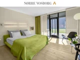 Moderne hotelværelse med en dobbeltseng dækket af grønt sengetøj, store vinduer med gennemsigtige gardiner, en stol og lyst trægulv. Sollyset strømmer ind i rummet.