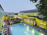 En stor udendørs swimmingpool ved siden af en lysegul bygning med liggestole, parasoller og en spiseplads på terrassen, omgivet af grønne områder i skumringen.