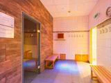 Et lyst, flisebelagt omklædningsrum med træbænke, vægkroge, et ur og en åben dør, der fører til en sauna beklædt med træpaneler.