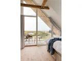 Et lyst soveværelse med trægulve, synlige bjælker, en dobbeltseng og store glasdøre ud til en balkon med en stol og naturskøn udendørs udsigt.