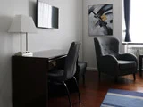 Et moderne værelse med et mørkt skrivebord, en lampe, to grå stole, et vægmonteret tv, et abstrakt maleri, et vindue med mørke gardiner og et blåt tæppe.