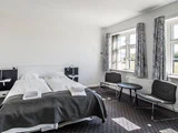 Lyst hotelværelse med to enkeltsenge skubbet sammen, gråt sengetøj, to stole, et lille rundt bord og store vinduer med mønstrede gardiner, der lukker naturligt lys ind.