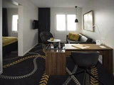 Moderne hotelværelse med mørkt mønstret gulvtæppe, skrivebord og stol i træ, sofa i gule nuancer, lænestol og stort vindue med gardiner, der lukker naturligt lys ind.