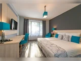 Moderne hotelværelse med en stor seng, blågrønne detaljer, skrivebord, stol, lænestol, fladskærms-tv og et vindue med gennemsigtige gardiner, der lukker naturligt lys ind.
