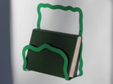En grøn, bølgekantet væghængt metalreol holder en mørkegrøn bog oprejst mod en hvid baggrund.