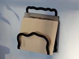 En vægmonteret magasinholder i sort metal med bølgede kanter holder en beige bog eller et magasin mod en lyseblå baggrund.