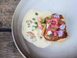 En tallerkenanrettet gourmetret med stegt fisk med rosa rogn, spiselige blomster og urter, serveret med cremet sauce, grøntsager i skiver og garniture på et rustikt træbord.