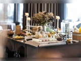 Et buffetbord med stearinlys, blomster, glas med yoghurt, en vanddispenser i glas, tallerkener med mad og elegant service i et blødt oplyst spiseområde.