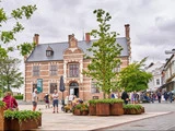 Folk går og samles på et livligt bytorv med moderne planter, træer og en historisk murstensbygning med gavle og et ur under en overskyet himmel.