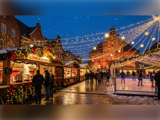Et festligt julemarked udendørs i skumringen med lys, folk, der slentrer rundt, madboder og en skøjtebane ved siden af pyntede, historiske murstensbygninger.