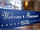 Et blåt skilt med hvid tekst, hvor der står Welcome Benvenuto og Hotel Mediterran, står ved en indgang.
