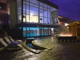 En moderne spa om natten med store glasvinduer, en indendørs pool, liggestole og to personer, der slapper af i et rundt udendørs boblebad.