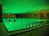 To personer svømmer i en stor indendørs pool med glødende grønt vand og et loft dekoreret med stjernelignende lys. Buede trin fører ned i poolen.