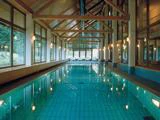 Indendørs swimmingpool med turkise fliser, træbjælker, store vinduer og liggestole i den fjerneste ende, omgivet af varm belysning og udsigt til træerne udenfor.