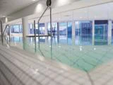 Indendørs swimmingpool med klart vand, flisebelagte kanter, metalgelændere og store vinduer, der lukker naturligt lys ind. Moderne, lyst og rent interiør.