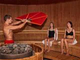 En mand vifter med en stor rød vifte i en træsauna, mens to kvinder i badetøj sidder på en bænk og nyder varmen.