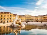 Barokpalads med gul facade og mange vinduer, bag et stort springvand med klassiske statuer og klart vand, under en blå himmel med tynde skyer.
