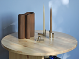 Et rundt træbord med en brun, bølget vase, to høje stearinlys i moderne stager og en lille cylindrisk genstand mod en lyseblå væg.
