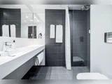 Moderne badeværelse med mørkegrå fliser, hvide bordplader, dobbelte håndvaske, et vægspejl, hvide håndklæder og en brusekabine med forhæng.