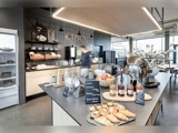 Moderne køkkencafé med kager, syltetøjsglas, frugt og morgenmadsprodukter på en disk. En person bevæger sig i nærheden af en kaffemaskine. Hylder, tallerkener og brød er synlige i baggrunden.