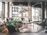 Moderne, solbeskinnet stue med store vinduer, grå sofaer, en gulvlampe, indendørs planter og sofaborde med udsigt til et urbant gadebillede udenfor.
