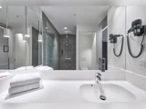 Moderne hotelbadeværelse med hvid håndvask, foldede håndklæder, væghængt hårtørrer, stort spejl og en glasindkapslet bruser med grå fliser i baggrunden.