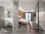 Moderne hotelværelse med dobbeltseng, skrivebord, stol, stort vindue med udsigt over byen og et badeværelse med håndvask og spejl adskilt af en delvis væg.