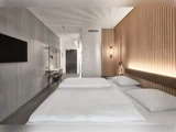 Moderne hotelværelse med minimalistisk indretning, en stor seng med hvidt sengetøj, et væghængt tv, et skrivebord og træpaneler på væggen. Varm belysning skaber en hyggelig atmosfære.