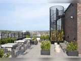 Tagterrasse med borde, stole og planter ved siden af en murstensvæg og en sort vindeltrappe med udsigt over et bybillede under en klar himmel.