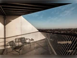 En moderne balkon med metalstole og et bord har udsigt over et bybillede under en blå himmel med stærke geometriske skygger og rækværk.