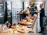 Et buffetbord med forskellige oste, frugter og grøntsager; en person serverer mad i baggrunden i en lys, moderne restaurant.