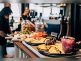 Et buffetbord med forskellige oste, vindruer, grønt og krukker med mad; folk serverer for sig selv i baggrunden i et varmt lys.