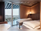 Et moderne hotelværelse med to enkeltsenge, trævægge, en væghængt lampe og en balkon med stole og et bord med udsigt over et udendørs område.