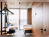 Minimalistisk hotelværelse med træmøbler, seng, skrivebord og åbent skab; et stort vindue fører ud til en balkon med stole og udsigt over byen.