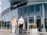 To personer med kufferter går mod glasindgangen til en moderne hotelbygning på en solrig dag. På hotelskiltet over døren står der A Place To Hotel.