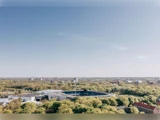 Luftfoto af et stort stadion omgivet af grønne træer og bygninger under en klar blå himmel.