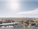En skarp sol skinner over et bybillede med røde murstensbygninger, grønne træer og fjerne industristrukturer under en klar himmel.