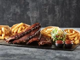 Et fad med grillede ribben, tre portioner pommes frites, en grøn salat i en glasskål og to kopper barbecuesauce på et skiferbræt.
